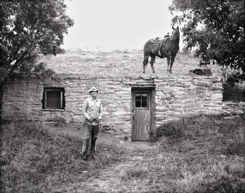 Frank Perry standing before a stone dugout house built in the late 1890s, a saddled horse on the roofline above.
